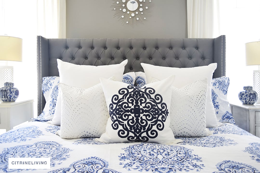 Master bedroom with gorgeous blue and white bedding, grey upholstered bed and white drapes.