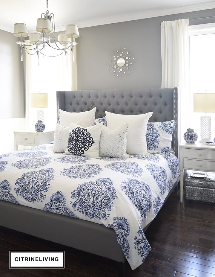 Master bedroom with gorgeous blue and white bedding, grey upholstered bed and white drapes.