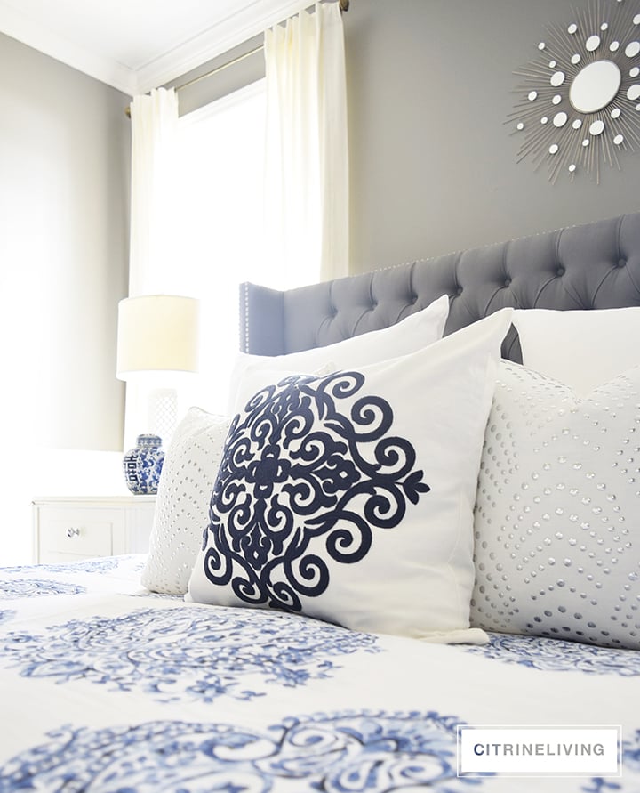 Master bedroom with gorgeous blue and white bedding, grey upholstered bed and white drapes.