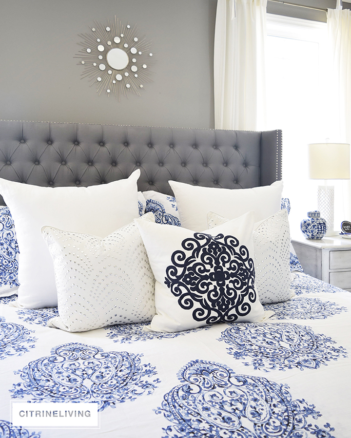 Master bedroom with gorgeous blue and white bedding, grey upholstered bed and white drapes.