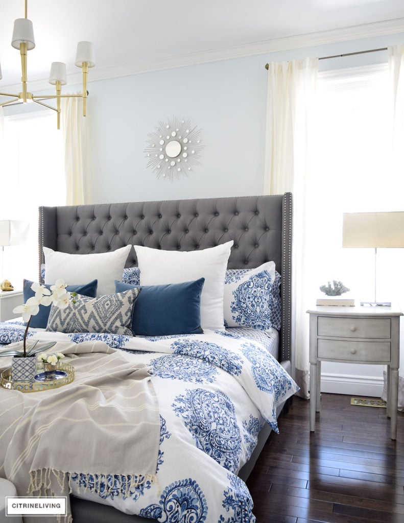 Gorgeous blue and white bedroom featuring blue and white bedding paired with global inspired textiles, grey upholstered bed and brass accents and lighting bring a bright and airy look to any bedroom. 