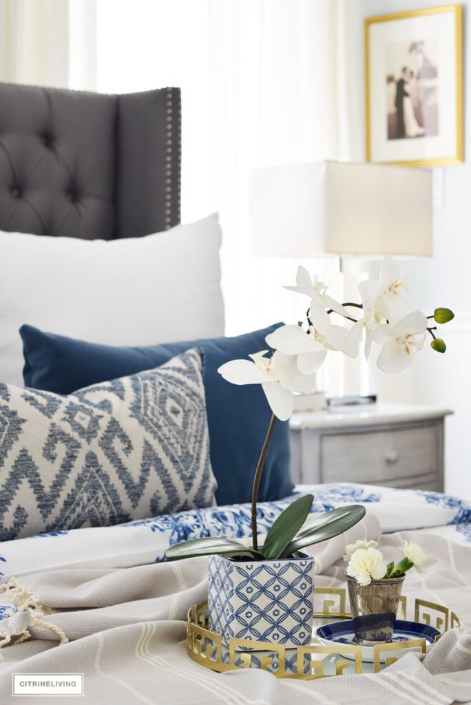 Gorgeous blue and white bedroom featuring blue and white bedding paired with global inspired textiles, grey upholstered bed and brass accents and lighting bring a bright and airy look to any bedroom. Layer your bed with small luxuries to create a true retreat - a gold mirrored tray and florals!