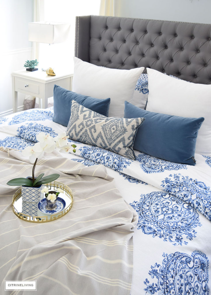 Gorgeous blue and white bedroom featuring blue and white bedding paired with global inspired textiles, grey upholstered bed and brass accents and lighting bring a bright and airy look to any bedroom. 