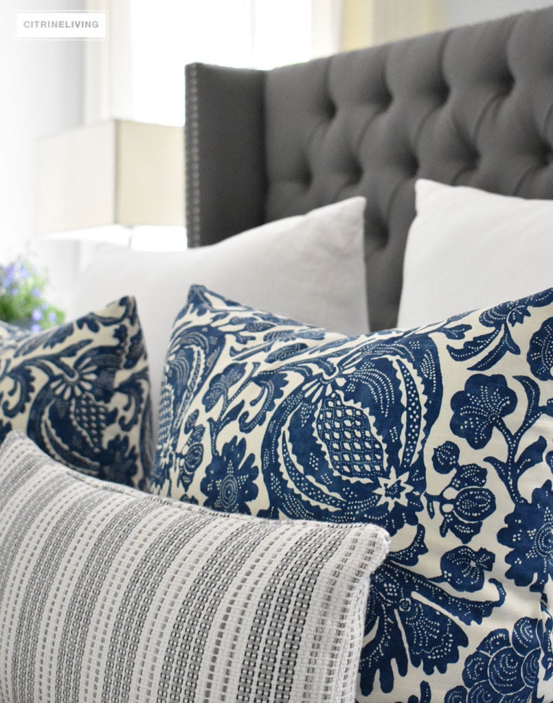 Blue and white Summer decorated bedroom with layers of bold pattern - batik, stripes and paisley - bring a casual, coastal look. Upholstered headboard with nailhead trim is classic and elegant. Light blue walls keep the look crisp and airy.