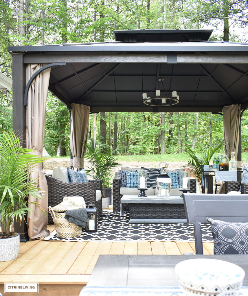 Create the perfect lounging area in your backyard with a beautiful covered gazebo, with added height for an airy, open feel. A gorgeous fire table, solar chandelier, resin wicker furniture, and layered accessories create an elegant and sophisticated look.