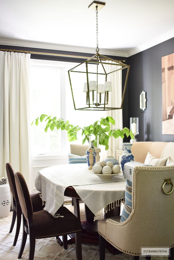 Sophisticated and elegant dining room with transom details. Black walls painted in Cracked pepper by Behr Paint. Neutral decor with layers of blue and white pottery. Upholstered chairs with antique brass nailed trim are elegant and refined. Large scale lantern pendant chandelier makes a bold statement. Tailored, custom white and grey pinstripe drapes and layers of striped textiles are classic touches.