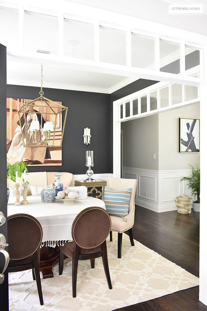 Sophisticated and elegant dining room with transom details. Black walls painted in Cracked pepper by Behr Paint. Neutral decor with layers of blue and white pottery. Upholstered chairs with antique brass nailed trim are elegant and refined. Large scale lantern pendant chandelier makes a bold statement.
