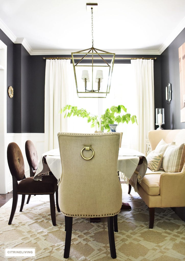 Sophisticated and elegant dining room with transom details. Black walls painted in Cracked pepper by Behr Paint. Neutral decor with layers of blue and white pottery. Upholstered chairs with antique brass nailed trim are elegant and refined. Large scale lantern pendant chandelier makes a bold statement. Tailored, custom white and grey pinstripe drapes and layers of striped textiles are classic touches.