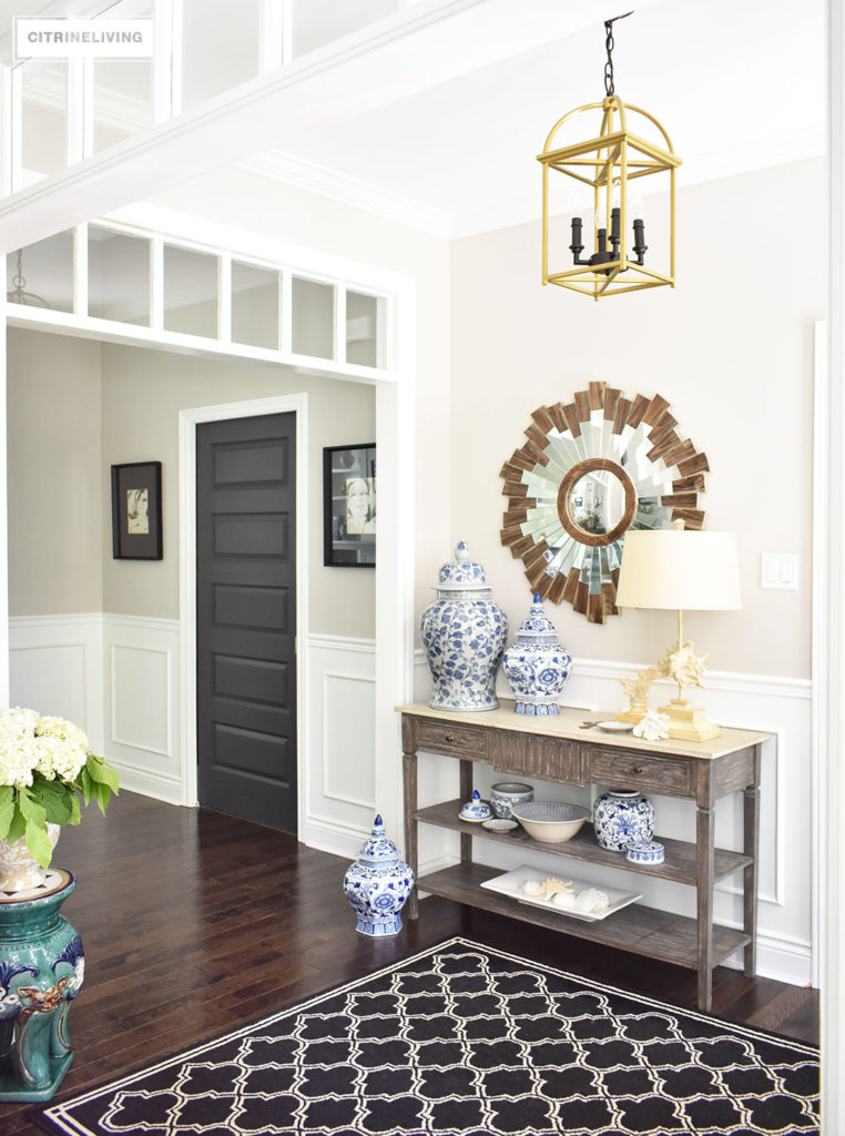 Large entryway with transom details lead to an open concept floor plan. Summer decorating with blue and white accents and fresh greenery keep the look light. Brass lantern style lighting and starburst mirror, a black and white graphic patterned rug help bring a sophisticated look.