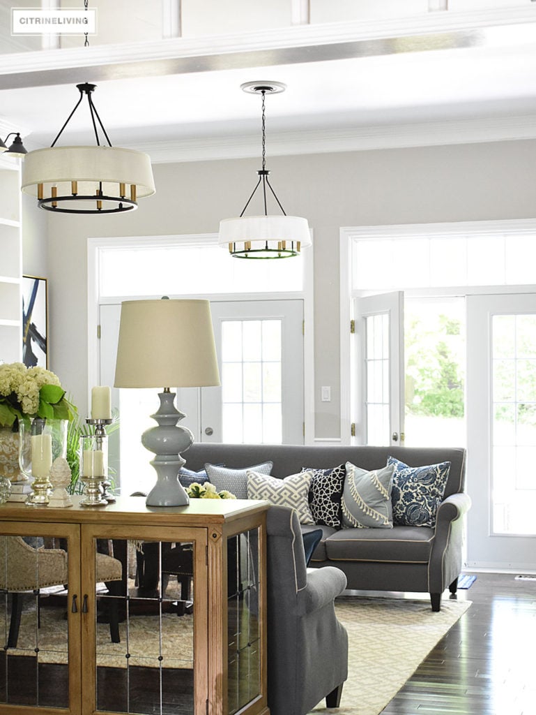 Elegant, open concept living room, with with grey sofas and drum shade pendant lights create a neutral backdrop of layers of beautiful blue patterned pillows. Decorated for Summer with fresh cut greenery, hydrangeas and palms. 