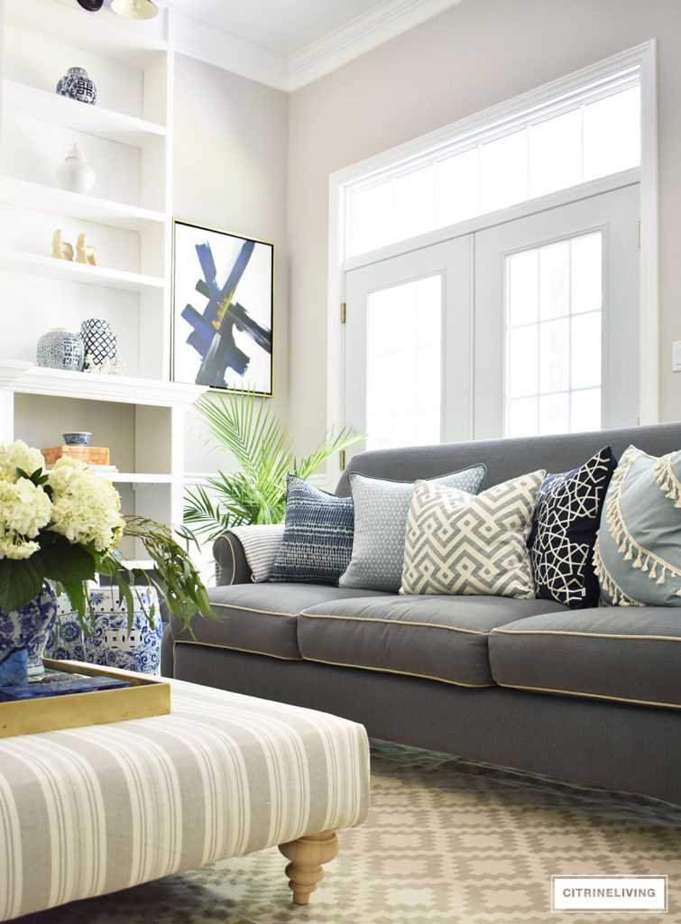Summer decorated living with layers of beautiful blue patterned pillows accented with fresh palms. Custom built bookshelves hold blue and white pieces mixed wit coastal accessories, brass accents and design books. A striped upholstered ottoman makes a classic statement.