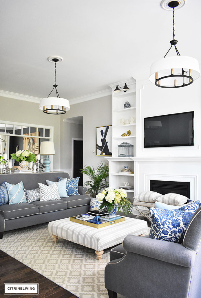 Elegant, open concept living room, with with grey sofas and drum shade pendant lights create a neutral backdrop of layers of beautiful blue patterned pillows and blue and white pottery. Decorated for Summer with fresh cut greenery, hydrangeas and palms. 
