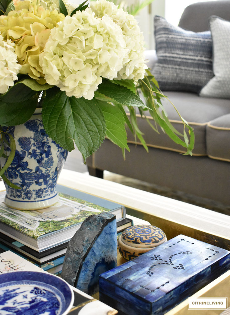 Summer decorated living with layers of beautiful blue patterned pillows, accented with fresh hydrangeas and greenery arranged in a blue an white ginger jar. Blue accessories and coastal elements enhance the Summer theme. 