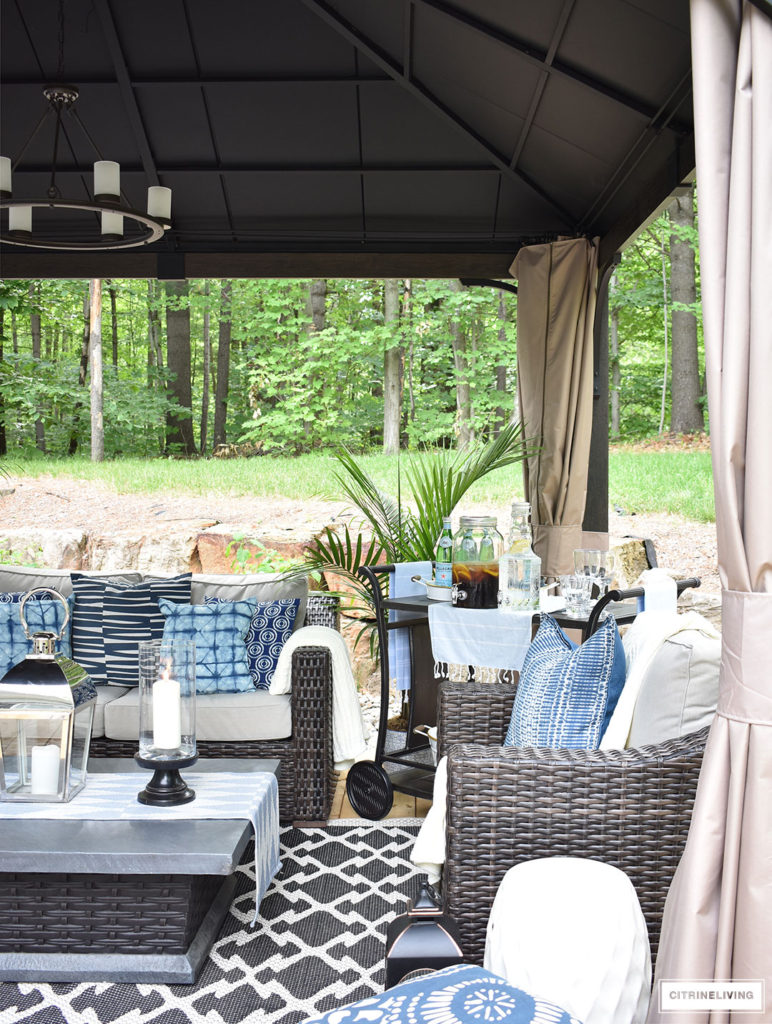 Create the perfect lounging area in your backyard with a beautiful covered gazebo, with added height for an airy, open feel. A gorgeous fire table, solar chandelier, resin wicker furniture, and layered accessories create an elegant and sophisticated look.