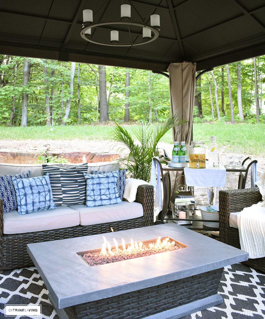 Create the perfect lounging area in your backyard with a beautiful covered gazebo, with added height for an airy, open feel. A gorgeous fire table to keep warm on chilly nights, solar chandelier, resin wicker furniture, and layered accessories create an elegant and sophisticated look.