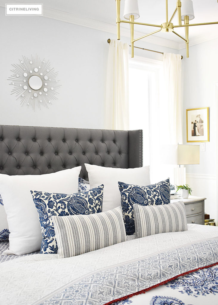 Blue and white Summer decorated bedroom with layers of bold pattern - batik, stripes and paisley - bring a casual, coastal look. Upholstered headboard with nailhead trim is classic and elegant. Light blue walls keep the look crisp and airy.