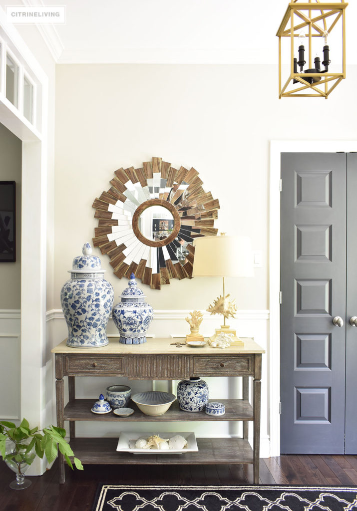 Large entryway with transom details lead to an open concept floor plan. Summer decorating with blue and white accents and fresh greenery keep the look light. Brass lantern style lighting and starburst mirror, a black and white graphic patterned rug help bring a sophisticated look.