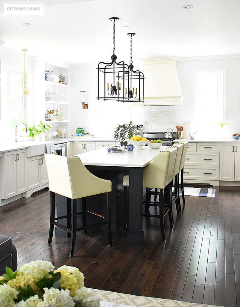 White kitchen with black island decorated for Summer with fresh cute greenery and bowls filled with bright, seasonal fresh fruit for a vibrant punch of seasonal color. Blue and white dishes and bowls are a classic touch. Lantern style pendant lighting over the island makes a bold statement.