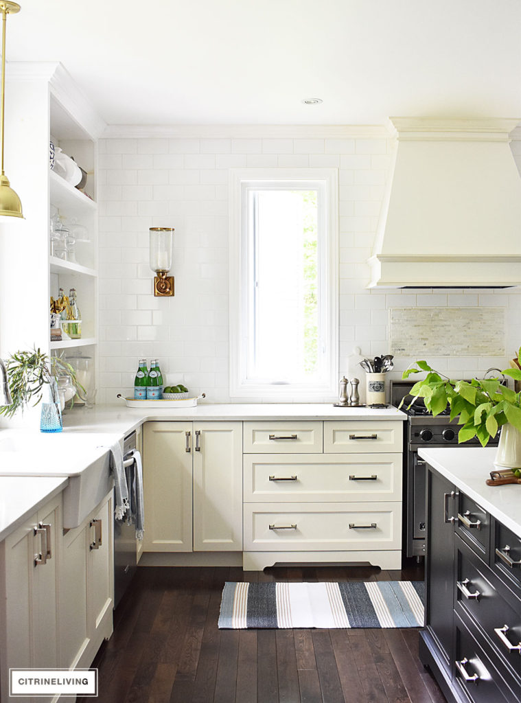 White kitchen with black island decorated for Summer with blue and white ginger jars and dishes accented a blue and white striped rug and turkish towels and fresh cute greenery.