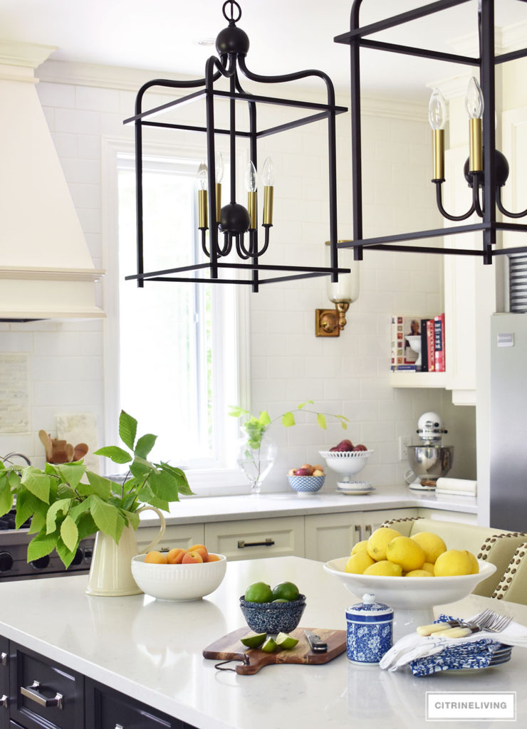 White kitchen with black island decorated for Summer with fresh cute greenery and bowls filled with bright, seasonal fresh fruit for a vibrant punch of seasonal color. Blue and white dishes and bowls are a classic touch. Lantern style pendant lighting over the island makes a bold statement.