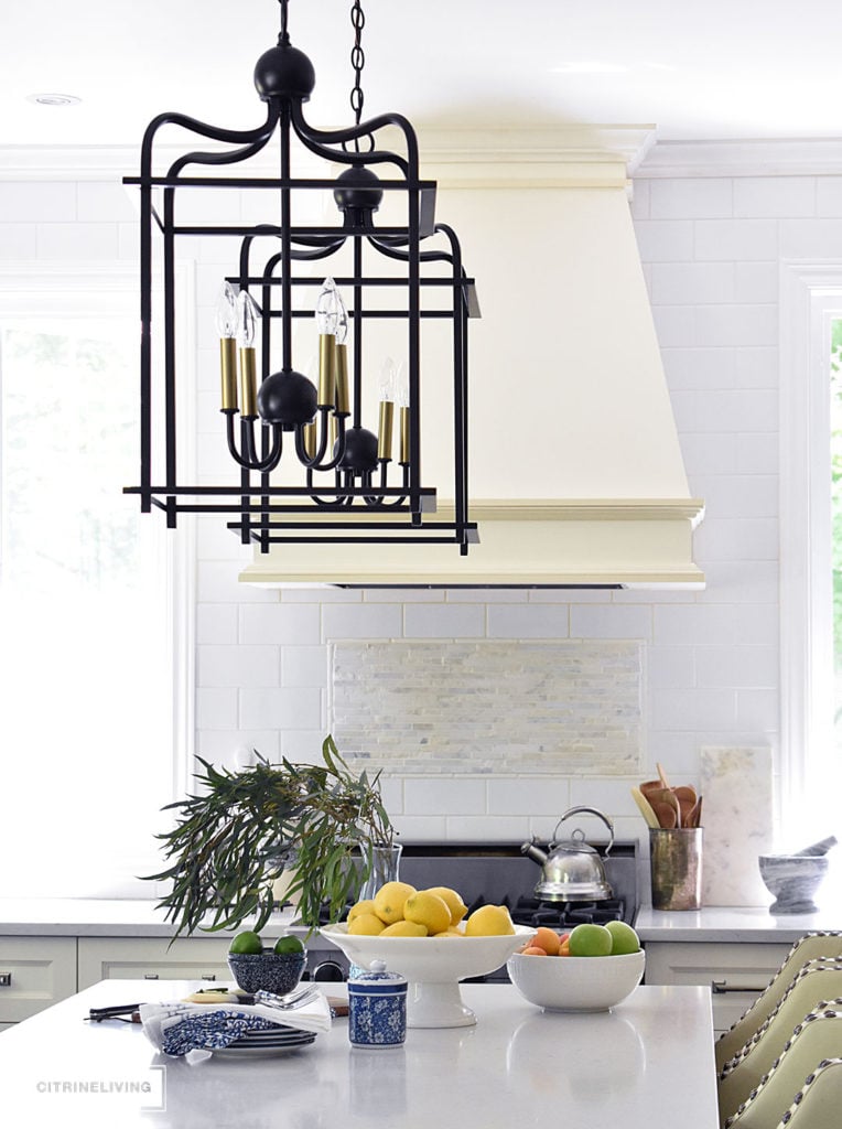 White kitchen with black island decorated for Summer with fresh cute greenery and bowls filled with bright, seasonal fresh fruit for a vibrant punch of seasonal color. Blue and white dishes and bowls are a classic touch. Lantern style pendant lighting over the island makes a bold statement.