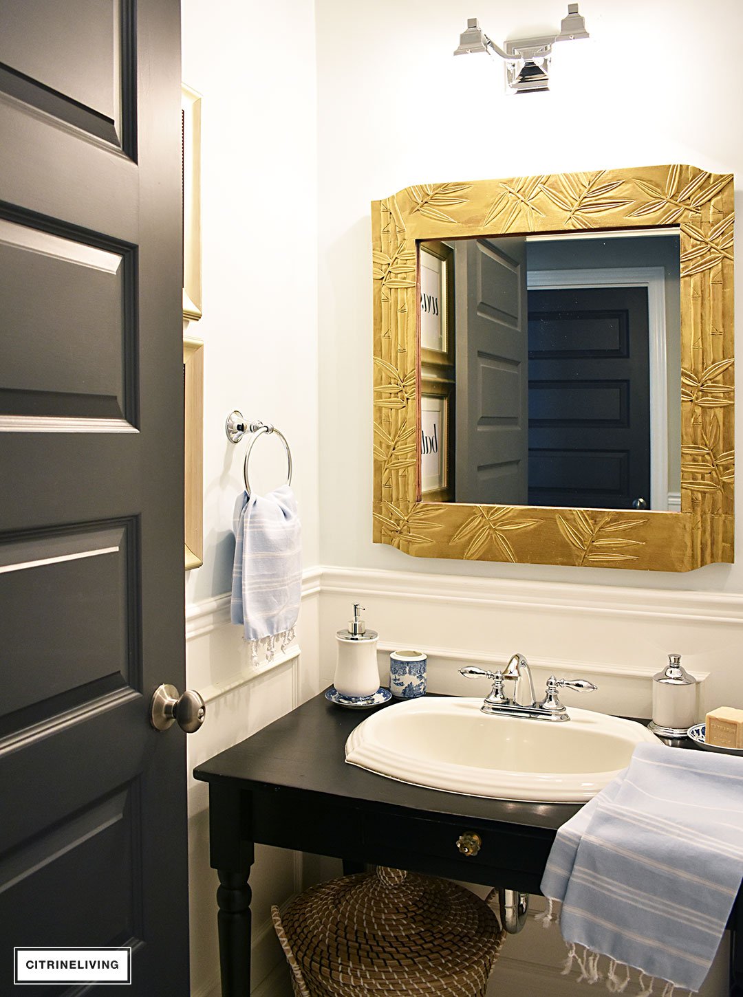 Small bathroom with white wainscotting, light blue walls, black vanity and gold accents.