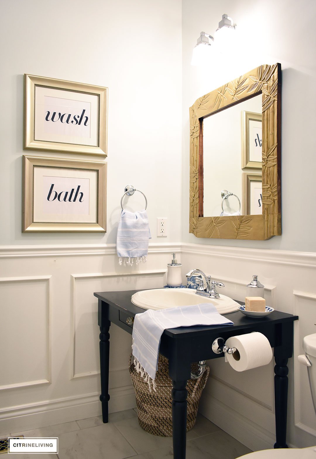 Small bathroom with white wainscotting, light blue walls, black vanity and gold accents.