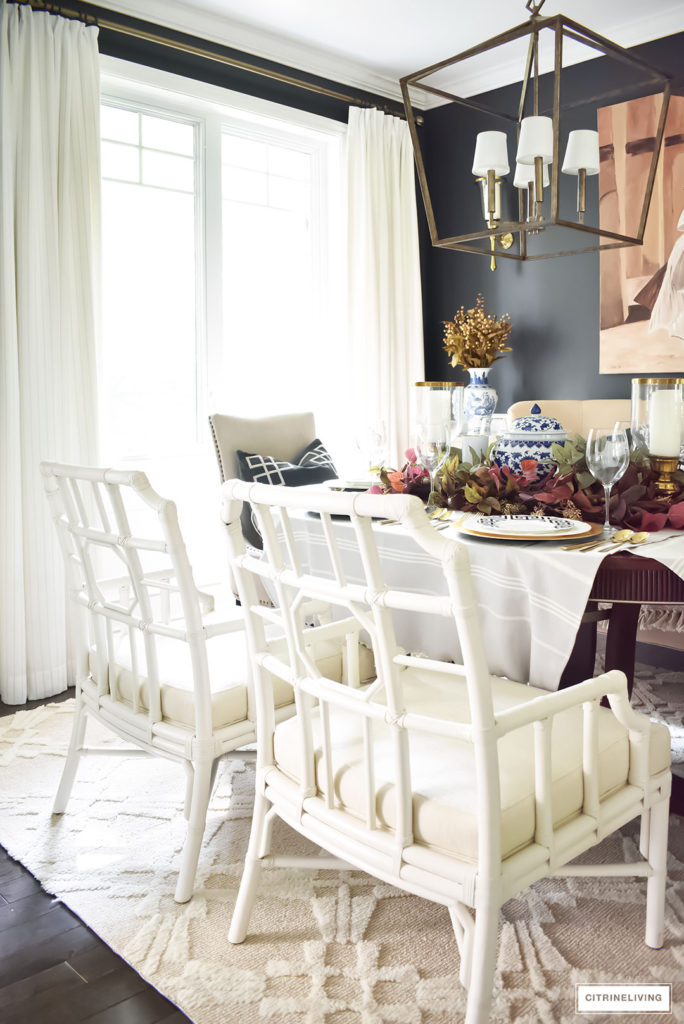 Dining room with black walls, white drapes, white lattice-back chairs, brass large scale chandelier. Fall tablescape with rich colors and blue and white accents.