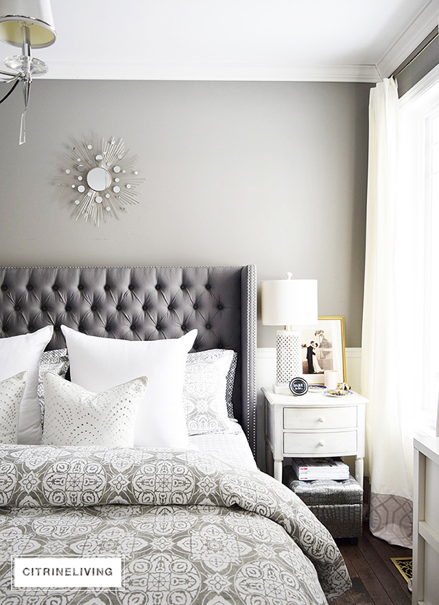 Beautiful master bedroom featuring upholstered headboard with nailhead trim and grey and white medallion print bedding. Ralph Lauren Washboard wall color.