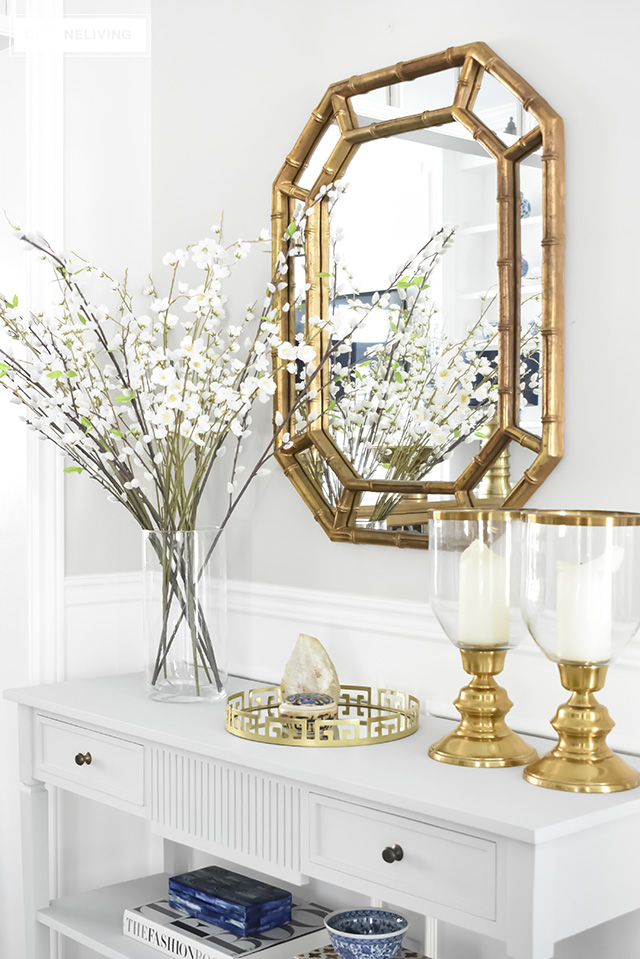Console table styled with gold and glass hurricane lantern, a gold tray and spring floral stems in a glass jar.