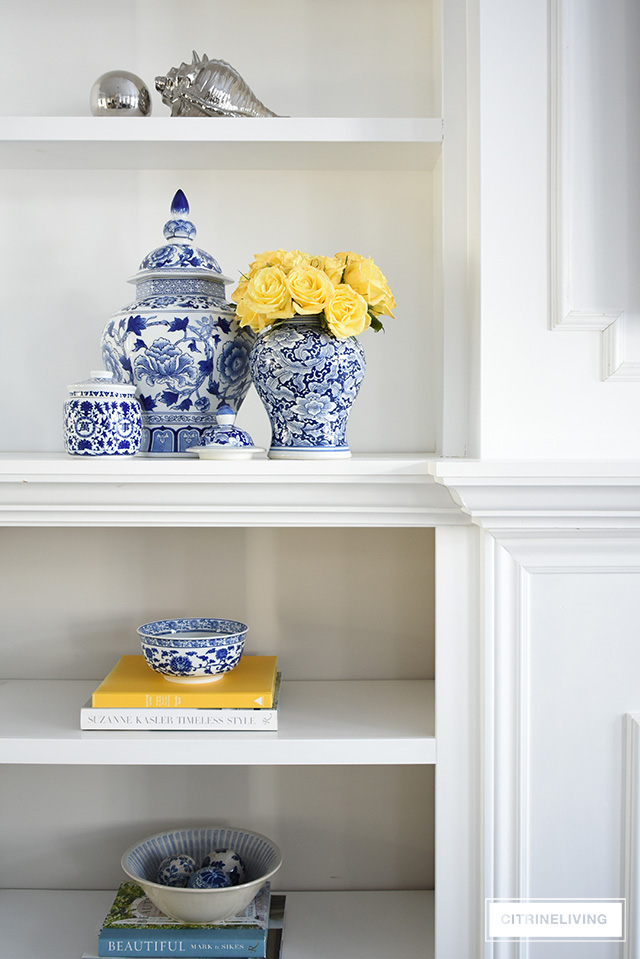 A beautifully decorated bookshelf with blue and white chinoiserie, ginger jars, yellow roses and decor accents.