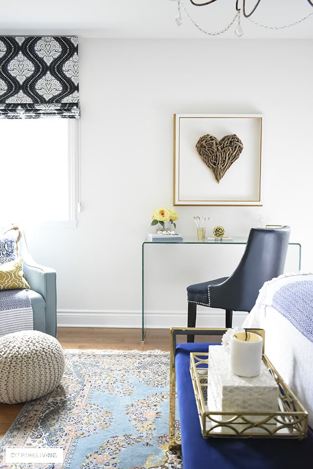 Beautiful writing desk area in this Boho-Glam bedroom featuring driftwood heart artwork paired with a modern waterfall glass desk. Layers of pattern and texture throughout the space add an eclectic, collected vibe.