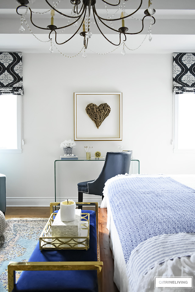 Beautiful writing desk area in this Boho-Glam bedroom featuring driftwood heart artwork paired with a modern waterfall glass desk. Layers of pattern and texture throughout the space add an eclectic, collected vibe.