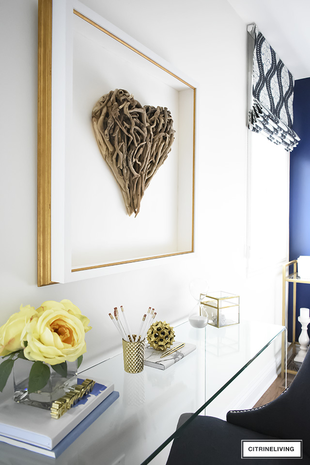 Beautiful writing desk area in this Boho-Glam bedroom featuring driftwood heart artwork paired with a modern waterfall glass desk. Layers of pattern and texture throughout the space add an eclectic, collected vibe.