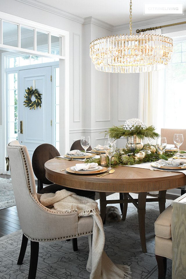 Christmas table with fresh greenery is perfect for holiday entertaining in this gorgeous dining room with elegant crystal chandelier, wall panelling and casual, relaxed furniture.