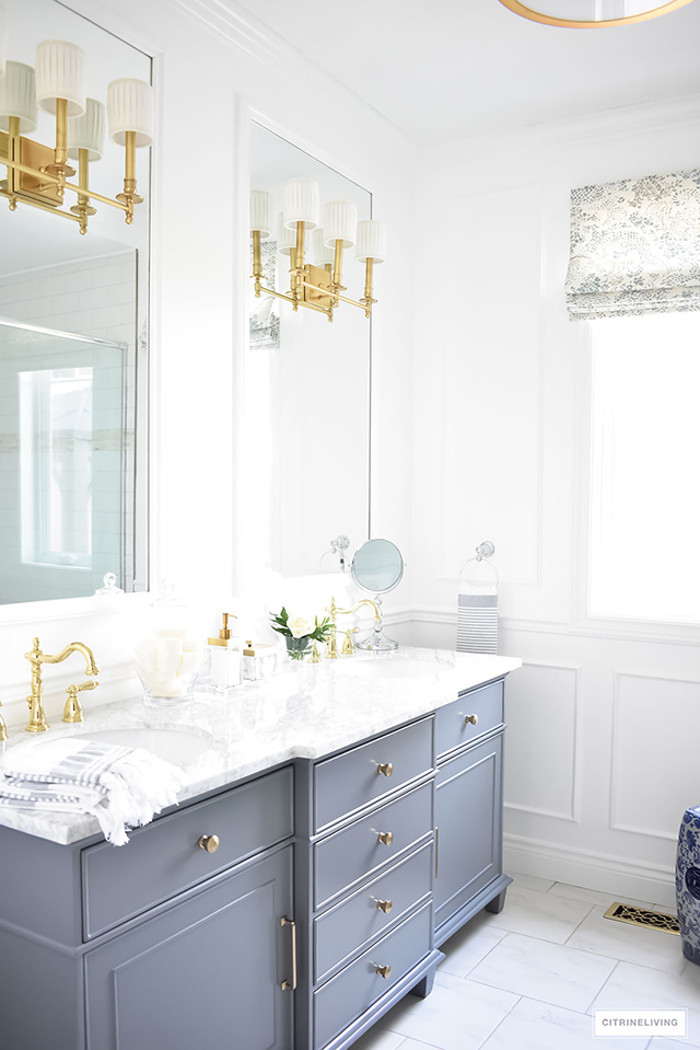White bathroom with elegant wall moldings and mirrors, brass wall sconces and a traditional grey vanity is sophisticated and lends a coastal look.