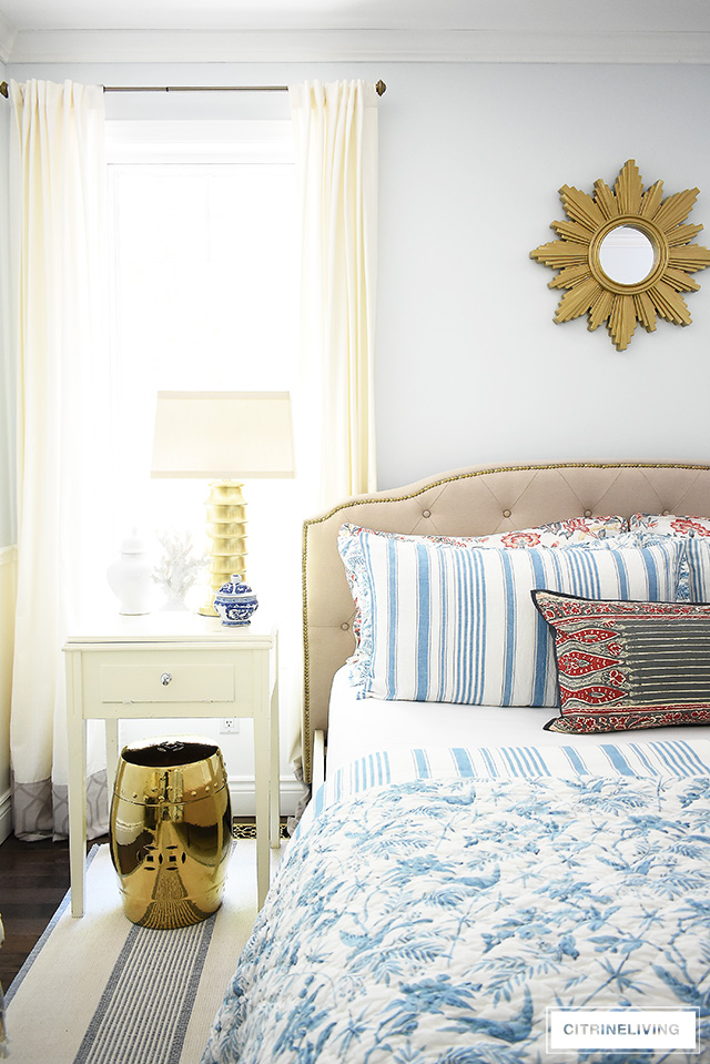 Gorgeous summer decorating in this bedroom featuring beautiful blue and white bedding mixed with red floras and block prints. Upholstered headboard with brass nailhead detail, brass lighting, starburst mirror and garden stool for a chic and elegant look.