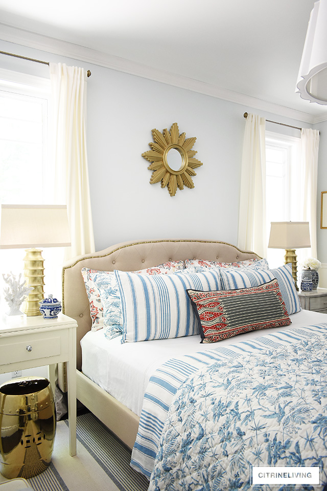 Traditional bedroom featuring a chic upholstered headboard with brass nailhead trim, brass lamps and starburst mirror. Summer bedding with a mix of blue and white stripes and red floral prints for a classic look.