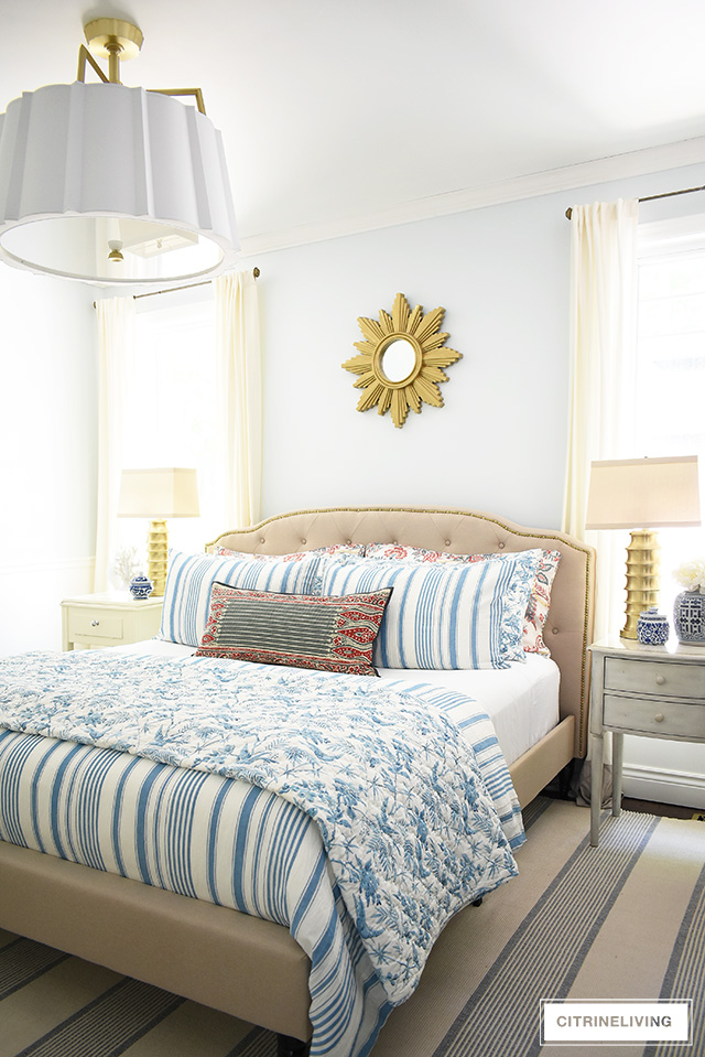 Beautiful summer decorated bedroom with blue and white striped bedding , upholstered bed with brass nailhead trim is chic and elegant. Over-scale drum shade chandelier with elegant brass detailing adds a chic and modern touch.