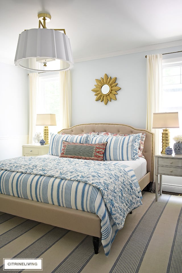 Beautiful summer decorated bedroom with blue and white striped bedding , upholstered bed with brass nailhead trim is chic and elegant. Over-scale drum shade chandelier with elegant brass detailing adds a chic and modern touch.