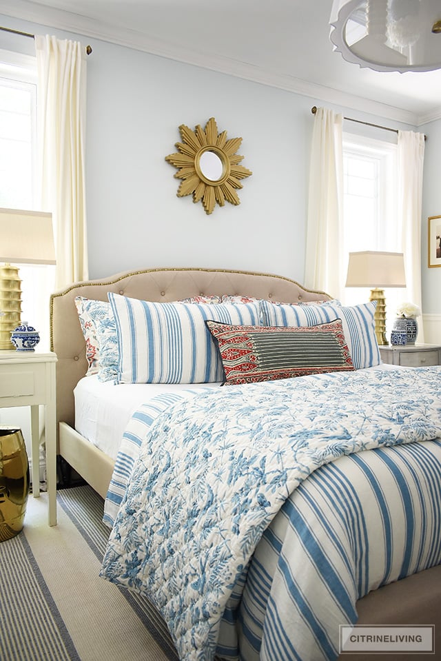 Traditional bedroom featuring a chic upholstered headboard with brass nailhead trim, brass lamps and starburst mirror. Summer bedding with a mix of blue and white stripes and red floral prints for a classic look.