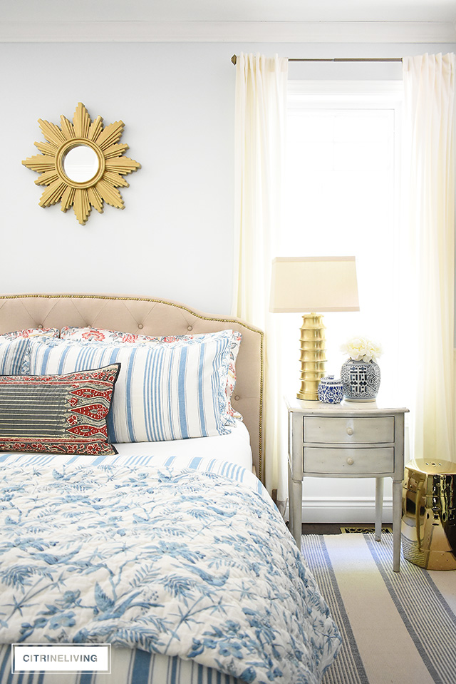Gorgeous summer decorating in this bedroom featuring beautiful blue and white bedding mixed with red floras and block prints. Upholstered headboard with brass nailhead detail, brass lighting, starburst mirror and garden stool for a chic and elegant look.