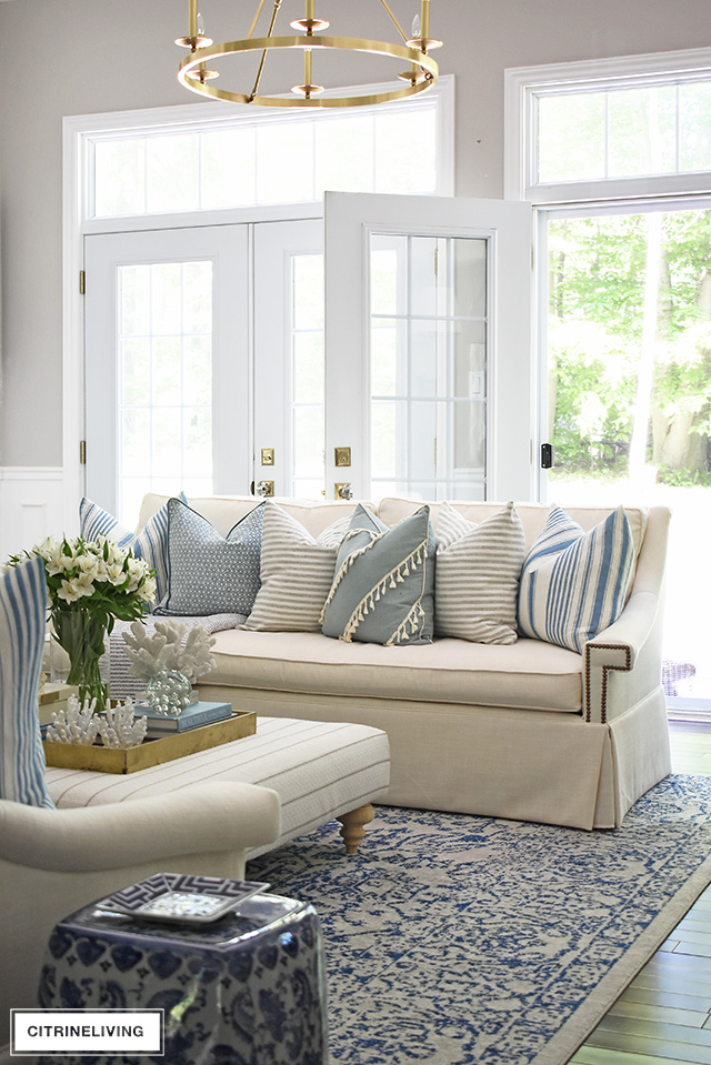 Bright and airy summer decorated living room featuring layered pillows with stripes and a light blue and white color palette for an elegant yet relaxed coastal theme.