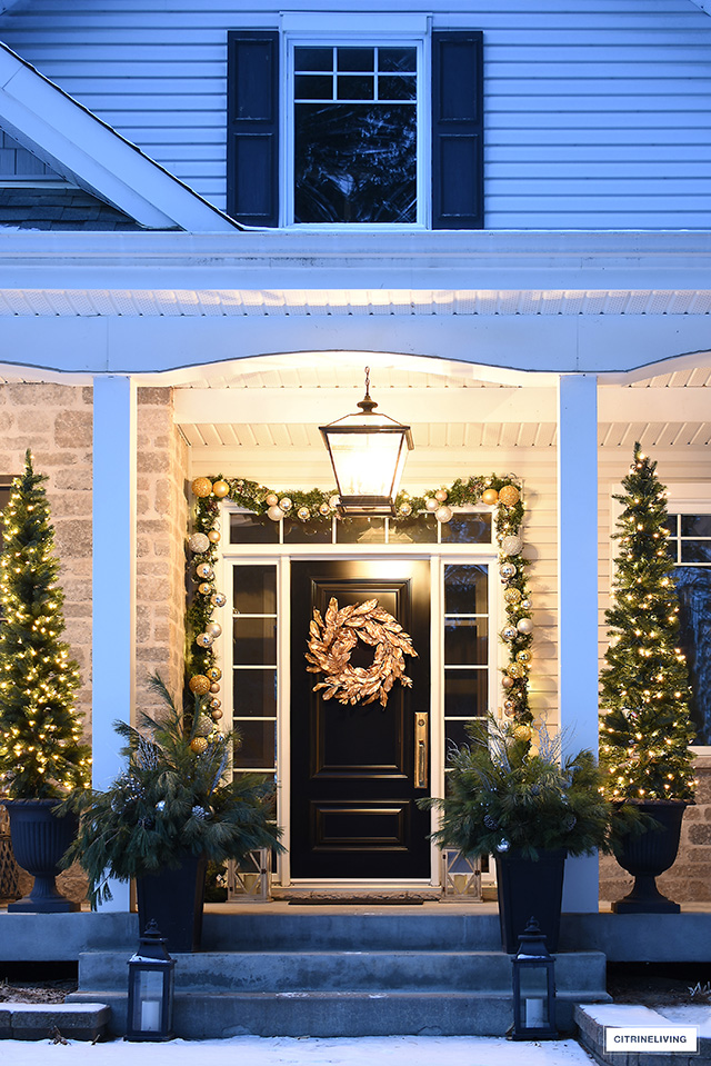 Elegantly decorated Christmas porch with lit pencil trees, garland and gold magnolia wreath.