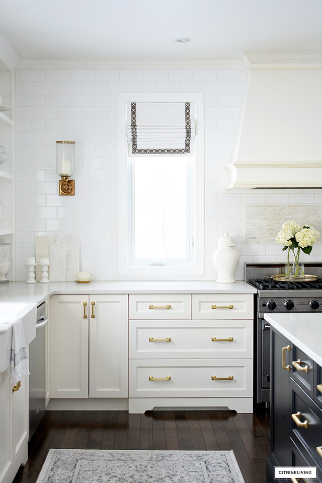White kitchen with brass hardware, custom rangehood, roman blinds on windows.