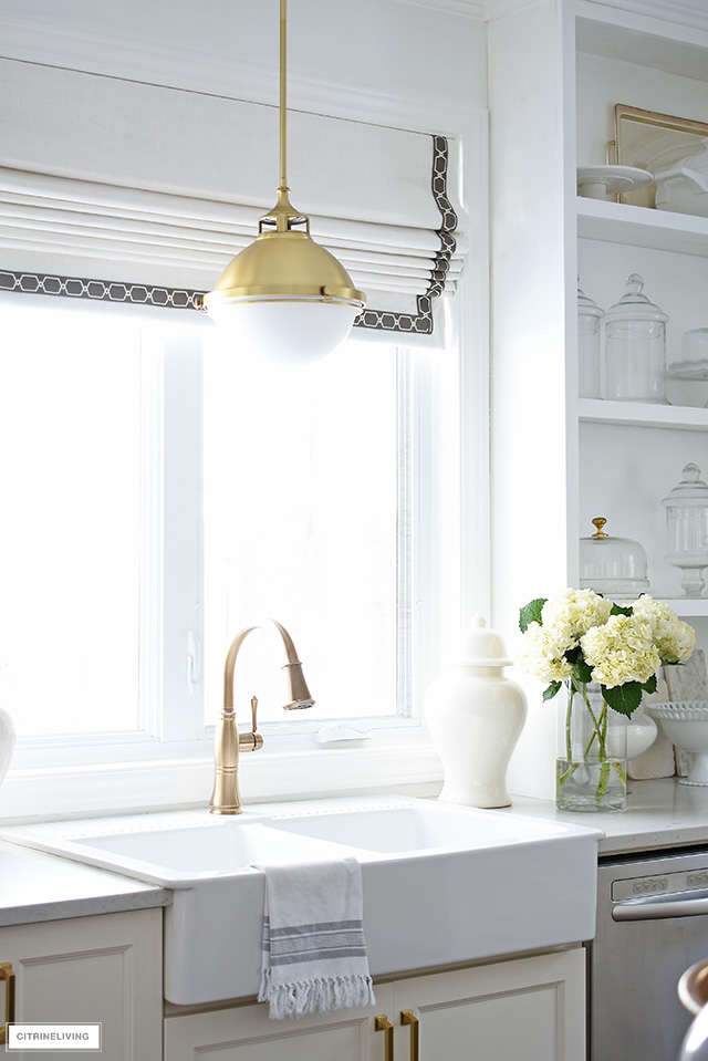 White farm sink with brass faucet and pendant light and custom Roman shade window covering.