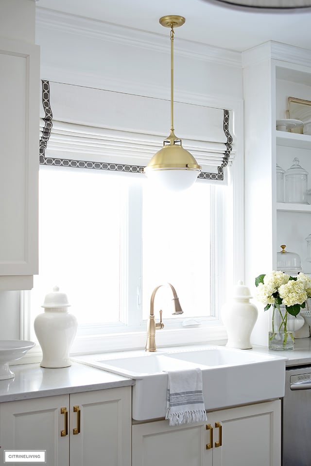 White farm sink with brass faucet and pendant light and custom Roman shade window covering.