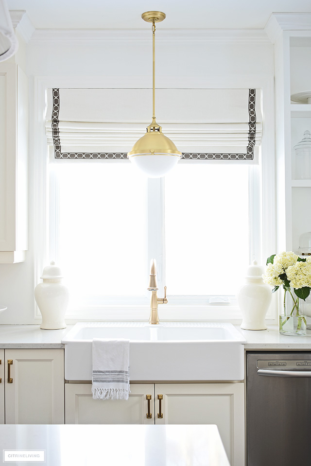 White farm sink with brass faucet and pendant light and custom Roman shade window covering.