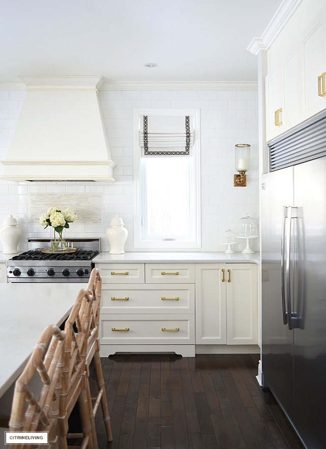 White kitchen with brass hardware, custom rangehood, roman blinds on windows.