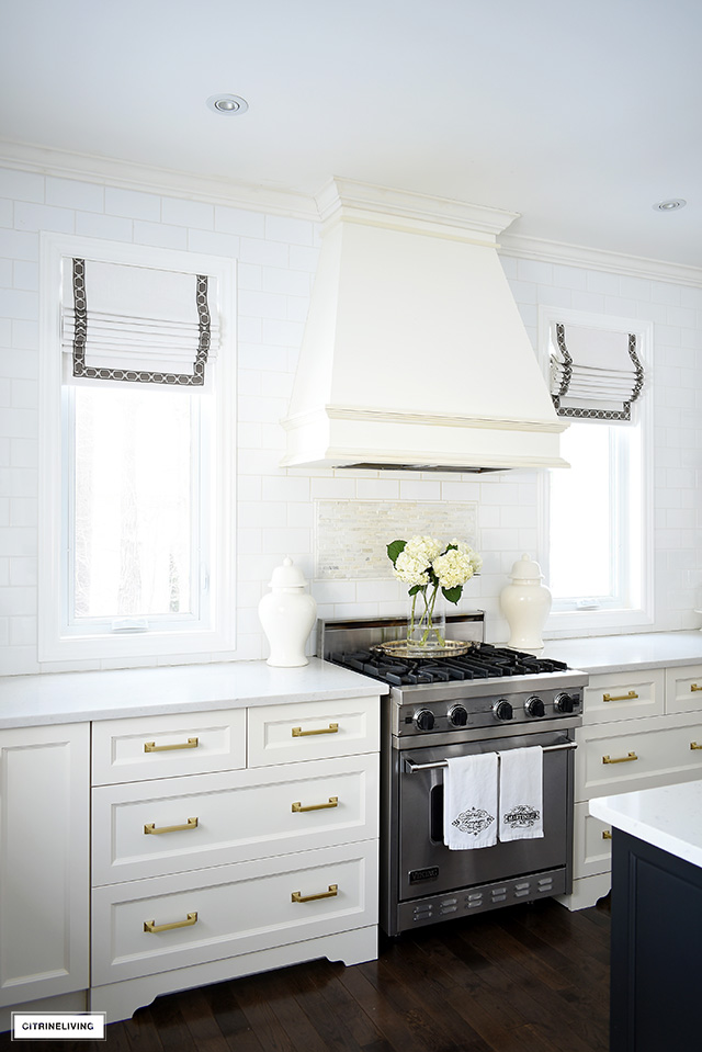 White kitchen with windows flanking the custom range hood, roman shades with ribbon detail.